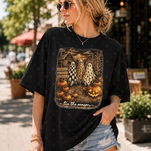 May include: A black short-sleeve t-shirt featuring a graphic print of three ghosts standing in a pumpkin patch near a barn. The print includes text reading PUMPKINS, HAY RIDES, CORN MAZE, and 'tis the season. The ghosts have checkered, leopard, and polka dot patterns.
