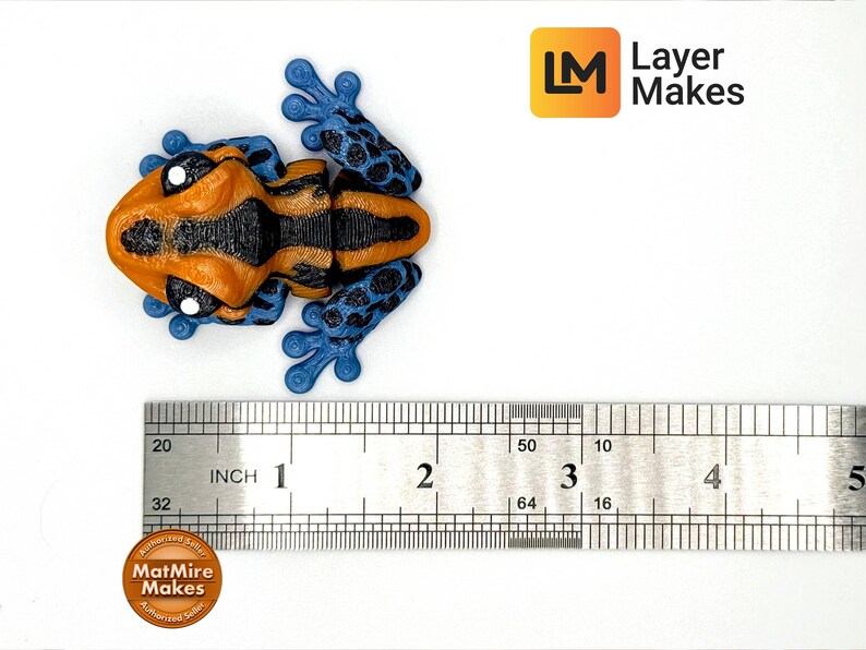 May include: A 3D-printed frog figurine in orange, black, and blue, with a ruler showing its size. The frog is approximately 3 inches long. The Layer Makes logo is visible in the upper right corner.