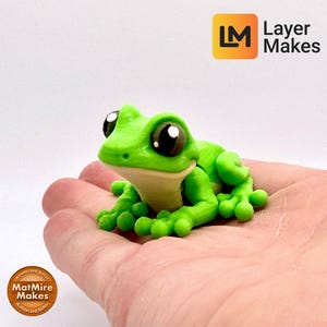 May include: A vibrant green frog figurine with large, expressive eyes and a light beige belly. The frog is resting on a hand, showcasing its detailed design. The Layer Makes logo is visible in the top right corner.