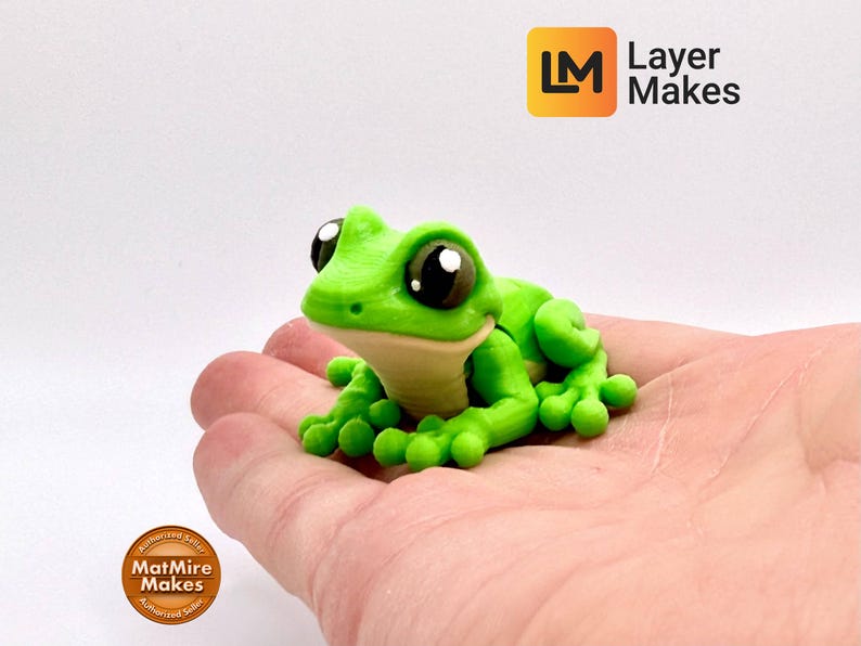 May include: A vibrant green frog figurine with large, expressive eyes and a light beige belly. The frog is resting on a hand, showcasing its detailed design. The Layer Makes logo is visible in the top right corner.