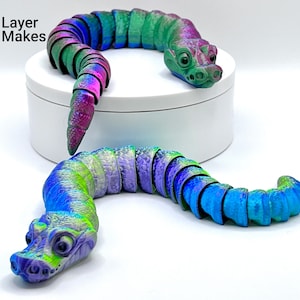 May include: Two iridescent, segmented, 3D printed snake-like figures with detailed heads. The snake on the left is green, blue, and purple. The snake on the right is pink, blue, and green.
