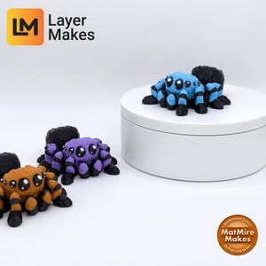 May include: Three colorful spider figurines. One is orange and black, another is purple and black, and the third is blue and black. The spiders have large, cartoonish eyes and are displayed on a white surface. The Layer Makes logo is in the top left.