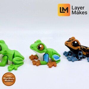 May include: Three colorful frog figurines. The first is green with white and brown accents. The second is green with blue and brown accents. The third is orange, black, and blue. The frogs are on a white surface.