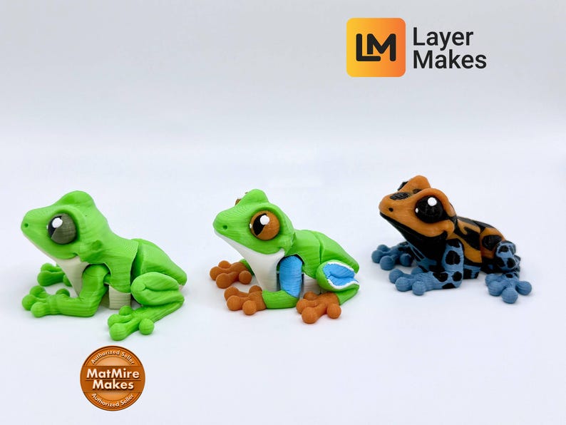 May include: Three colorful frog figurines. The first is green with white and brown accents. The second is green with blue and brown accents. The third is orange, black, and blue. The frogs are on a white surface.