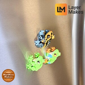 May include: Three colorful frog magnets. One frog is green, another is yellow and black, and the third is blue and black. The magnets are attached to a metallic surface. The Layer Makes logo is in the top right corner.