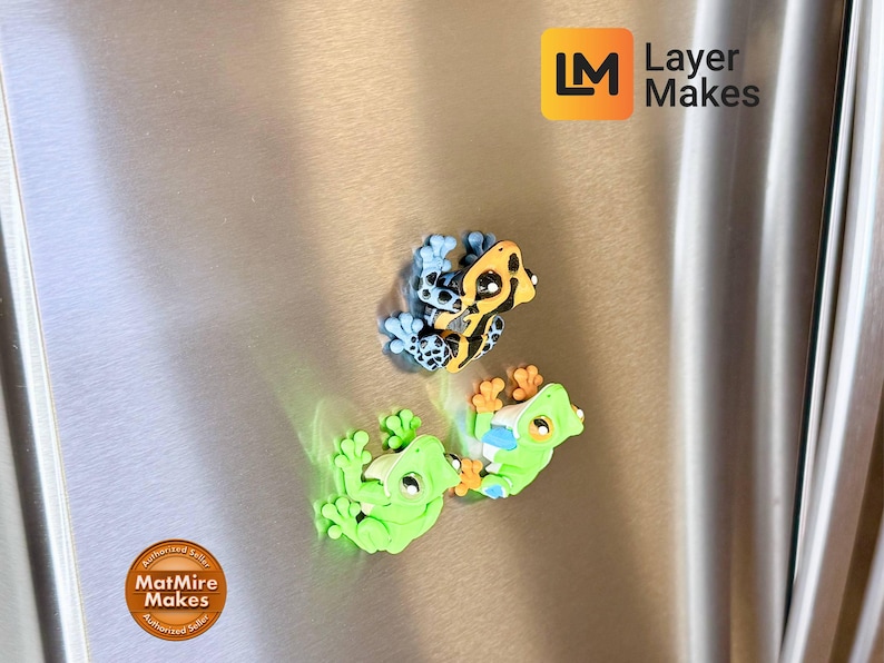 May include: Three colorful frog magnets. One frog is green, another is yellow and black, and the third is blue and black. The magnets are attached to a metallic surface. The Layer Makes logo is in the top right corner.