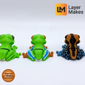 May include: Three colorful frog figurines in a row. The frogs are green, green with blue and orange, and orange and black with blue spots. The frogs are sitting on a white surface. The Layer Makes logo is in the top right corner.
