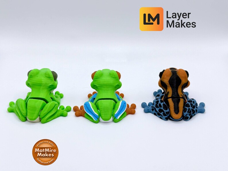 May include: Three colorful frog figurines in a row. The frogs are green, green with blue and orange, and orange and black with blue spots. The frogs are sitting on a white surface. The Layer Makes logo is in the top right corner.