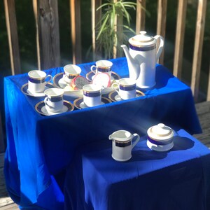 May include: A white porcelain tea set with blue and gold trim. The set includes a teapot, creamer, sugar bowl, and six cups and saucers. The set is displayed on a blue tablecloth.