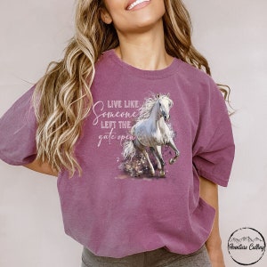 Peut inclure: T-shirt mauve avec un motif de cheval blanc en mouvement. Le texte "LIVE LIKE SOMEONE LEFT THE GATE OPEN" est au-dessus. Le vêtement a une coupe ample et des manches longues.