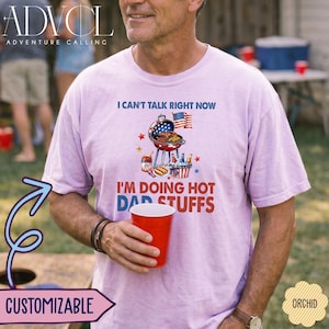 May include: A light purple short-sleeve t-shirt featuring a graphic of a charcoal grill, an American flag, and food items. The text reads I CAN'T TALK RIGHT NOW I'M DOING HOT DAD STUFFS in blue and red lettering. The shirt is worn by a person holding a red cup.