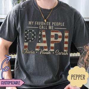 May include: Dark gray t-shirt with the text "MY FAVORITE PEOPLE CALL ME PAPI" in a patriotic design. The word "PAPI" is in red and blue, with the names Olivia, Noah, and Sarah below. The shirt is customizable.