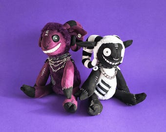 May include: Two plush ram dolls with button eyes and wide stitched grins. The left doll is purple with black horns and a spiked collar with chains. The right doll is black and white with striped horns and a spiked collar with a silver chain.