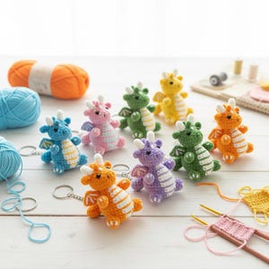 Może przedstawiać: Kolekcja kolorowych, szydełkowych breloków w kształcie smoka w różnych kolorach, w tym niebieskim, różowym, zielonym, żółtym, pomarańczowym i fioletowym. Każdy smok ma skrzydła i kółko do kluczy. W tle przędza i akcesoria do szydełkowania.