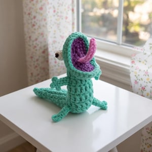 May include: A handmade crochet monster toy in shades of teal, purple, and pink. The creature has an open mouth with a long pink tongue, small arms, and a tail. It is sitting on a white surface.