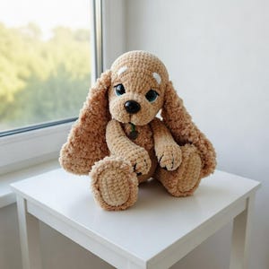 May include: A handmade, plush, tan-colored crochet dog with long floppy ears, blue eyes, and a black nose. The dog is sitting on a white table near a window. The dog has a small green tag around its neck.