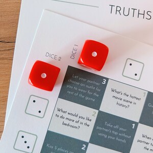 May include: A game board with a grid of 9 squares, each with a different truth or dare prompt. The prompts are written in white text on a dark gray background. Two red dice are on the board, one on the square labeled "DICE 1" and the other on the square labeled "DICE 2".