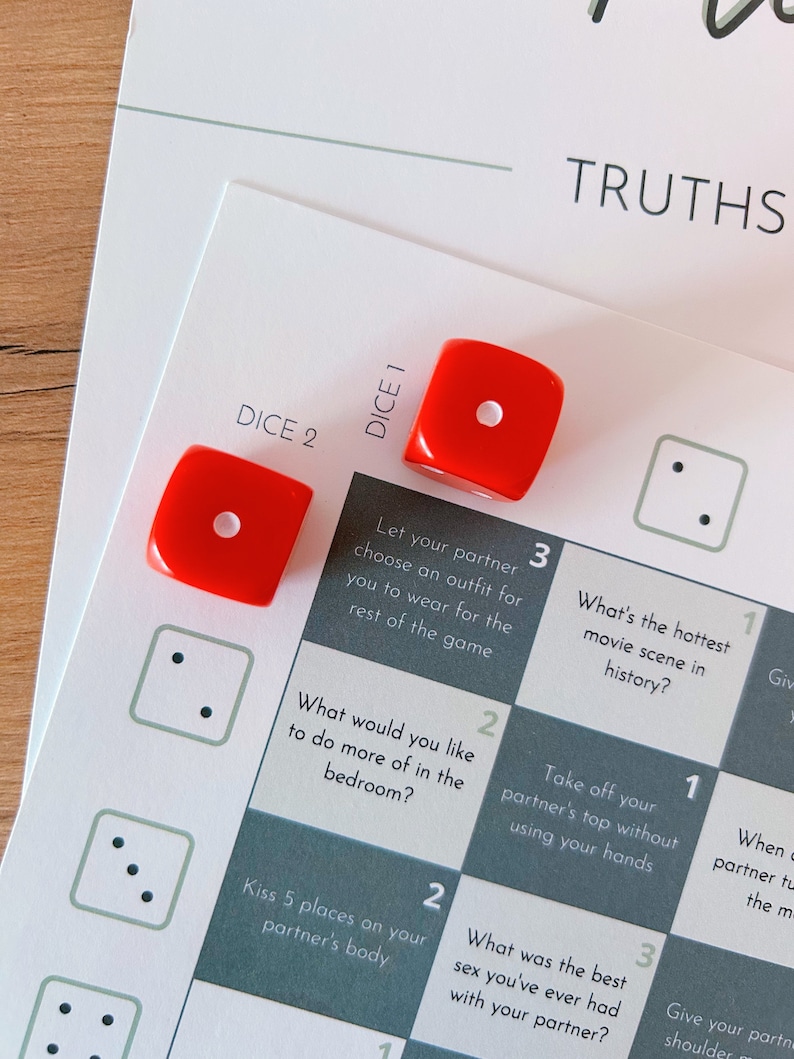 May include: A game board with a grid of 9 squares, each with a different truth or dare prompt. The prompts are written in white text on a dark gray background. Two red dice are on the board, one on the square labeled "DICE 1" and the other on the square labeled "DICE 2".