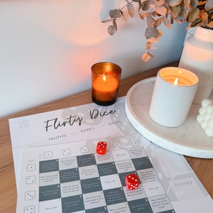 May include: A flirty dice game with a gray and white board, red dice, and instructions for truths, dares, and shots. The game is set on a wooden table with two candles and a plant.