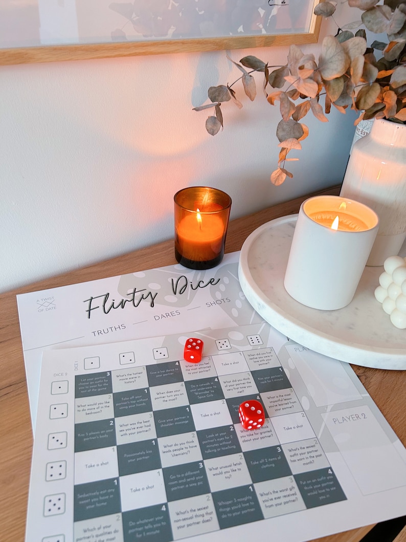 May include: A flirty dice game with a gray and white board, red dice, and instructions for truths, dares, and shots. The game is set on a wooden table with two candles and a plant.
