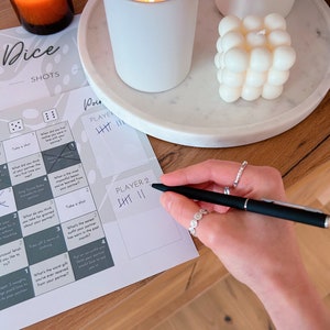 May include: A game board with dice and instructions for a drinking game called "Dice Shots". The board has a grid with different challenges and prompts for players to complete. The game is designed for two players and includes a scoring system. The board is on a wooden table with candles and a decorative object.