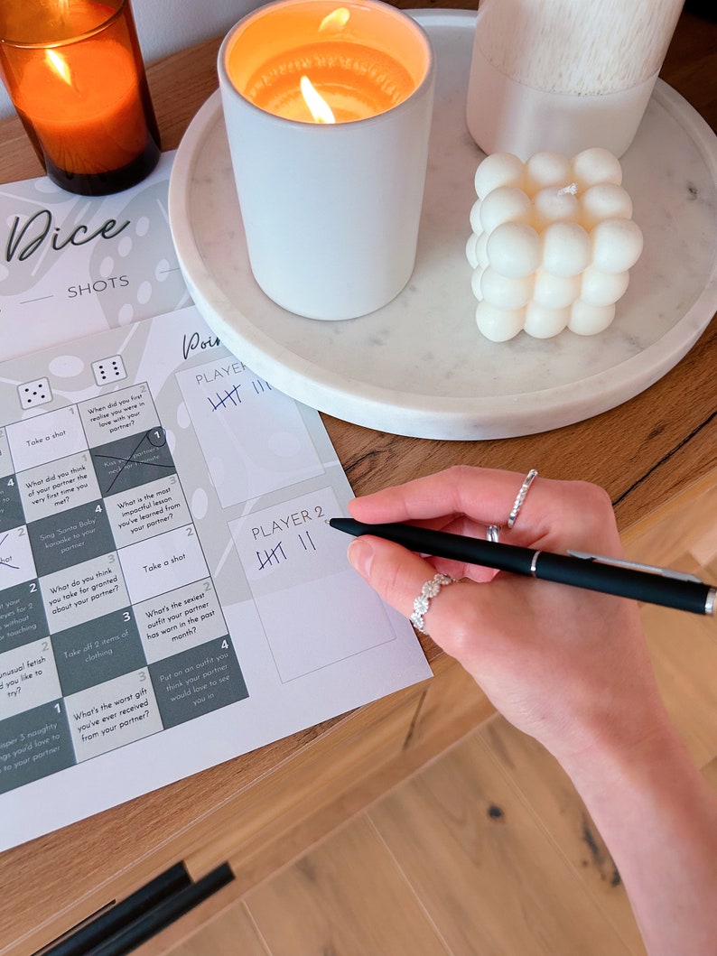 May include: A game board with dice and instructions for a drinking game called "Dice Shots". The board has a grid with different challenges and prompts for players to complete. The game is designed for two players and includes a scoring system. The board is on a wooden table with candles and a decorative object.