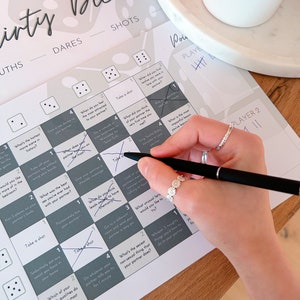 May include: A game board with a gray and white design. The board is divided into squares with numbers and prompts for a game called "Dirty Dice". The prompts include dares and truths. A hand is holding a pen and marking off a square on the board.