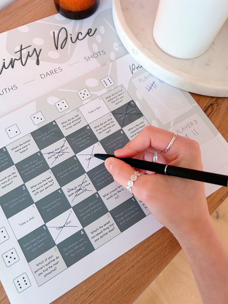 May include: A game board with a gray and white design. The board is divided into squares with numbers and prompts for a game called "Dirty Dice". The prompts include dares and truths. A hand is holding a pen and marking off a square on the board.