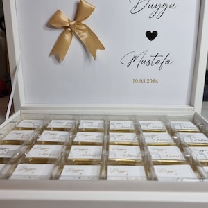 Könnte beinhalten: Eine weiße rechteckige Geschenkbox mit mehreren kleinen, einzeln verpackten goldfarbenen Gegenständen. Der Innendeckel ist mit einer goldenen Schleife und dem Text Duygu & Mustafa sowie einem Herz und dem Datum 10.05.2024 versehen.