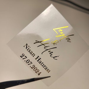 Puede incluir: Una pegatina cuadrada blanca con letras doradas. El texto dice "Leyla & Hilmi Nisan Hatırası 27.07.2024".