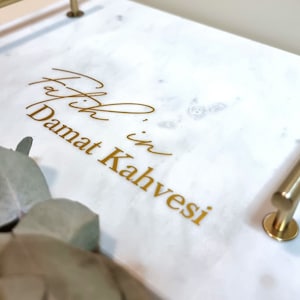 Pode incluir: Uma bandeja de servir de mármore branco com alças douradas e a inscrição "Fatih in Damat Kahvesi" em letras douradas.