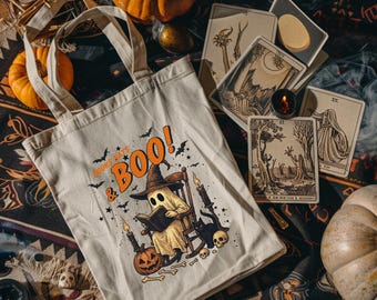 Puede incluir: Bolso tote beige con un diseño de Halloween. El diseño presenta un fantasma leyendo un libro, un gato negro y el texto "Books & Boo!". La escena incluye calabazas, velas y cartas del tarot.