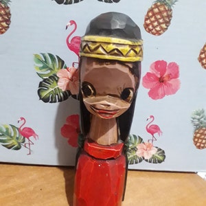 Op de afbeelding: Een houten beeldje van een persoon met lang zwart haar, gekleed in een rode jurk en een geel hoofdband. Het beeldje heeft een gegraveerd gezicht met grote ogen en een kleine mond.