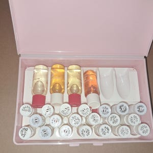 May include: A pink plastic case containing several small glass bottles with colored liquids and white caps. The bottles have red, white, and clear caps. The case also holds several white nail tips and additional white-capped bottles.