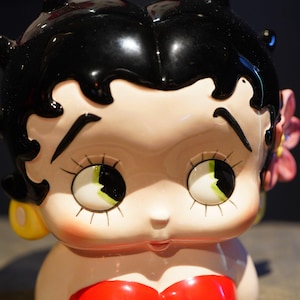 Könnte beinhalten: Eine Keramikfigur einer Zeichentrickfigur mit glänzendem schwarzem Haar, großen Augen mit langen Wimpern und einer kleinen Nase. Die Figur trägt gelbe Ohrringe und eine rosa Blume hinter einem Ohr sowie ein rotes Kleidungsstück.