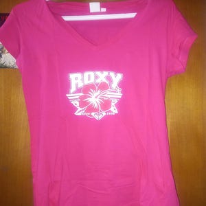 May include: Pink t-shirt with a white Roxy logo and a white hibiscus flower graphic. The logo says "Roxy" and "Since 1990".