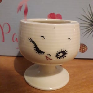 Può includere: Posacenere in ceramica bianca con un viso stilizzato con ciglia e una bocca rossa. Il posacenere ha la forma di un calice con una base rotonda.