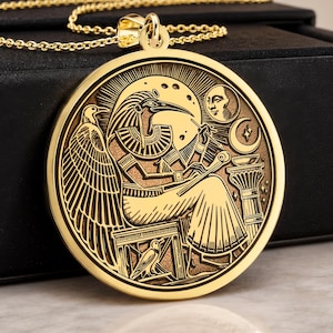 May include: A gold-toned circular pendant necklace featuring an engraved depiction of the Egyptian deity Thoth seated with a stylus and tablet, accompanied by a crescent moon and sun, suspended from a delicate matching gold-toned chain.