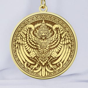 Op de afbeelding: Een goudkleurige hangerketting met een gedetailleerde cirkelvormige medaillon. De medaillon toont een gedetailleerd reliëf van een mythisch gevleugeld wezen met ingewikkelde patronen. De hanger hangt aan een delicate gouden ketting.