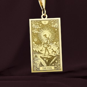 以下が含まれることがあります： 「愚者」のカードの画像が描かれたタロットカードのデザインが施された、ゴールドのペンダントネックレス。カードは、金色の縁取りが施された長方形で、崖の上に犬と一緒に立っている人物の細密なイラストが描かれています。