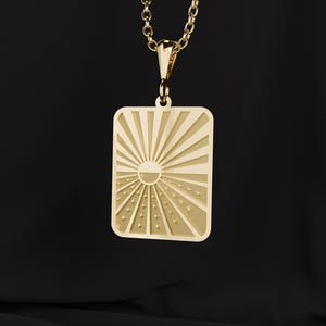 以下が含まれることがあります： 長方形のペンダントが付いたゴールドカラーのネックレス。ペンダントは、太陽と放射線、小さな星のようなディテールを備えたサンバーストデザインが特徴です。ネックレスは繊細なゴールドチェーンから吊り下げられています。