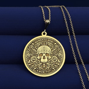 Gold tone Memento Mori necklace on dark blue fabric with an engraved skull, roses, hourglasses, scrolling leaves, and Amor Fati inscription.