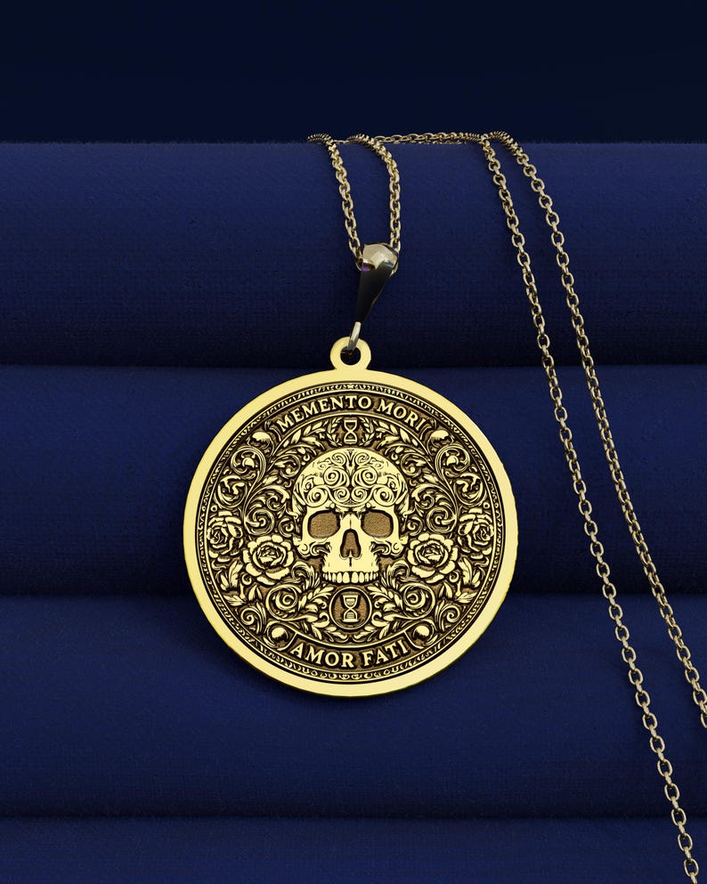 Gold tone Memento Mori necklace on dark blue fabric with an engraved skull, roses, hourglasses, scrolling leaves, and Amor Fati inscription.