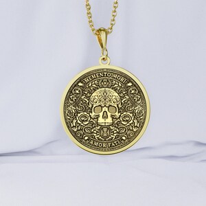 Gold tone Memento Mori necklace on white fabric with an engraved skull, roses, hourglasses, scrolling leaves, and Amor Fati inscription.