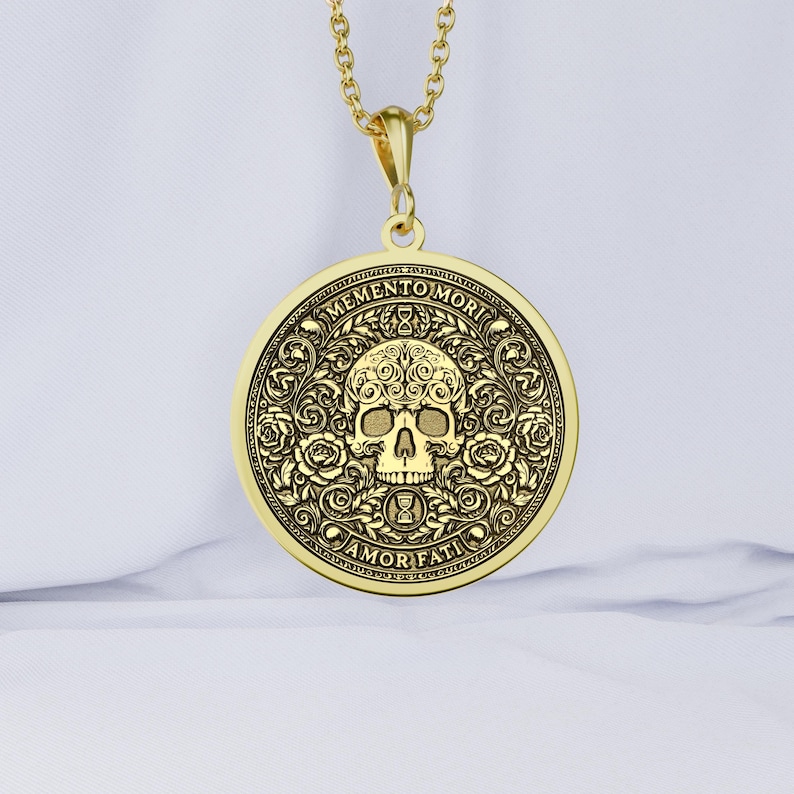 Gold tone Memento Mori necklace on white fabric with an engraved skull, roses, hourglasses, scrolling leaves, and Amor Fati inscription.