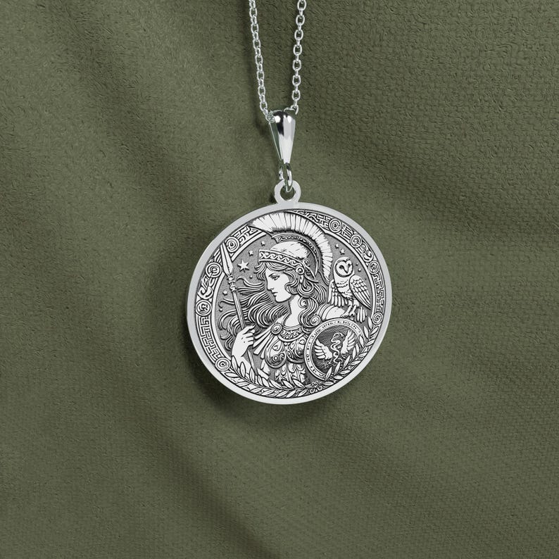 May include: Silver necklace with a round pendant featuring a detailed engraving of a woman in a helmet holding a spear, with an owl perched on her shoulder. The pendant has a Greek-inspired design.