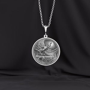 May include: A silver pendant necklace featuring a circular pendant with a detailed relief of a winged figure reaching towards a sun. The pendant is suspended from a delicate chain. The design includes architectural elements and waves.
