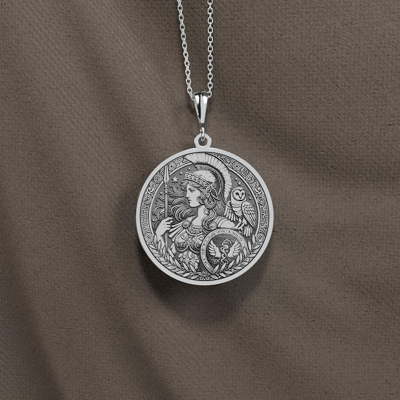 May include: Silver necklace with a round pendant featuring a detailed engraving of a woman in a helmet with an owl perched on her shoulder. The pendant has an inscription around the edge.