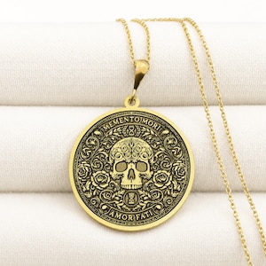 Gold tone Memento Mori necklace on cream fabric with an engraved skull, roses, hourglasses, scrolling leaves, and Amor Fati inscription.