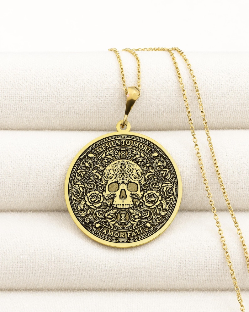 Gold tone Memento Mori necklace on cream fabric with an engraved skull, roses, hourglasses, scrolling leaves, and Amor Fati inscription.
